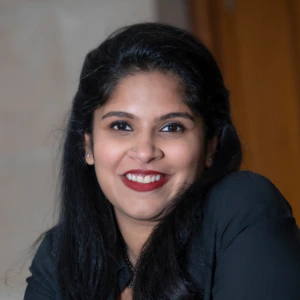 A woman with long dark hair, wearing a black top, smiles at the camera against a softly blurred indoor background.