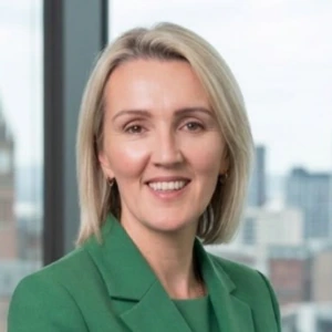 A woman with blonde hair, wearing a green blazer, stands smiling in an office with large windows and a cityscape in the background.