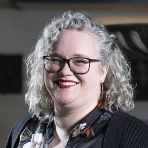 A woman with curly gray hair and glasses smiles, wearing a dark floral blouse and black cardigan, standing indoors with soft lighting.