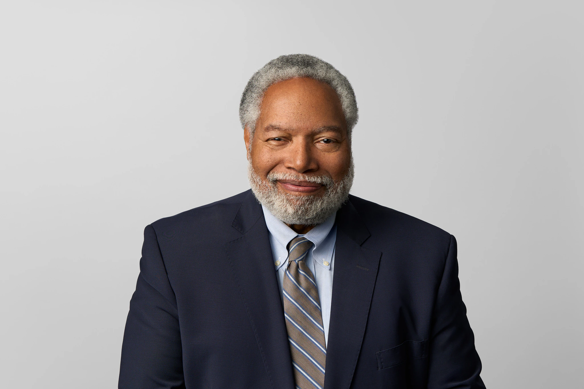 Smiling older man with gray hair and beard wearing a navy suit, light blue shirt, and striped tie, posing against a plain light gray background.