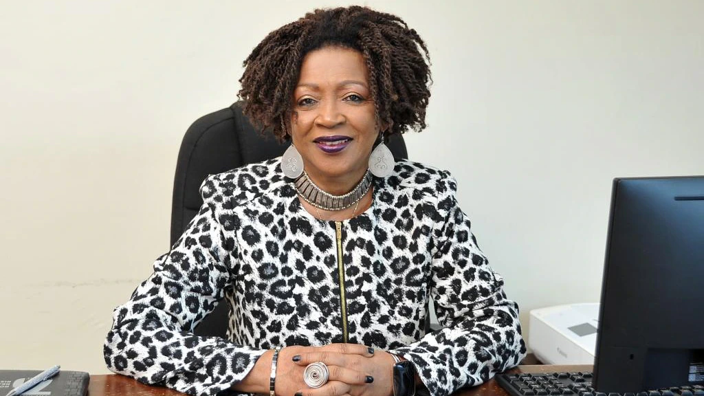 A woman with curly hair and statement jewelry sits at a desk, smiling. She wears a black and white patterned jacket. A computer and printer are on the desk.