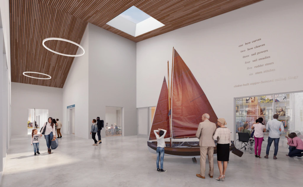 People stand in a bright, modern lobby with a wooden ceiling and skylight, surrounding a sailboat display. Some read a wall text, while others talk or look around. Circular lights hang from the ceiling.