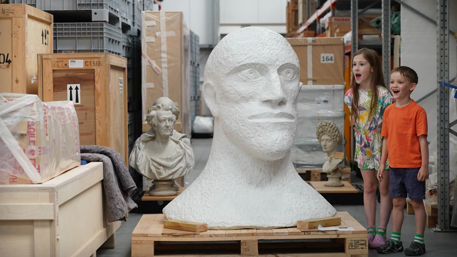 Two children stand next to a large white head sculpture in a storage area filled with crates, boxes, and classical busts. Both children appear excited and surprised.