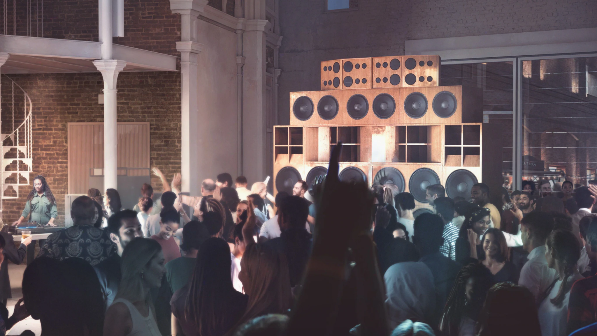 A crowded indoor party with people dancing in front of large stacked speakers. Light beams shine on the crowd, and a person stands by a spiral staircase in the background.