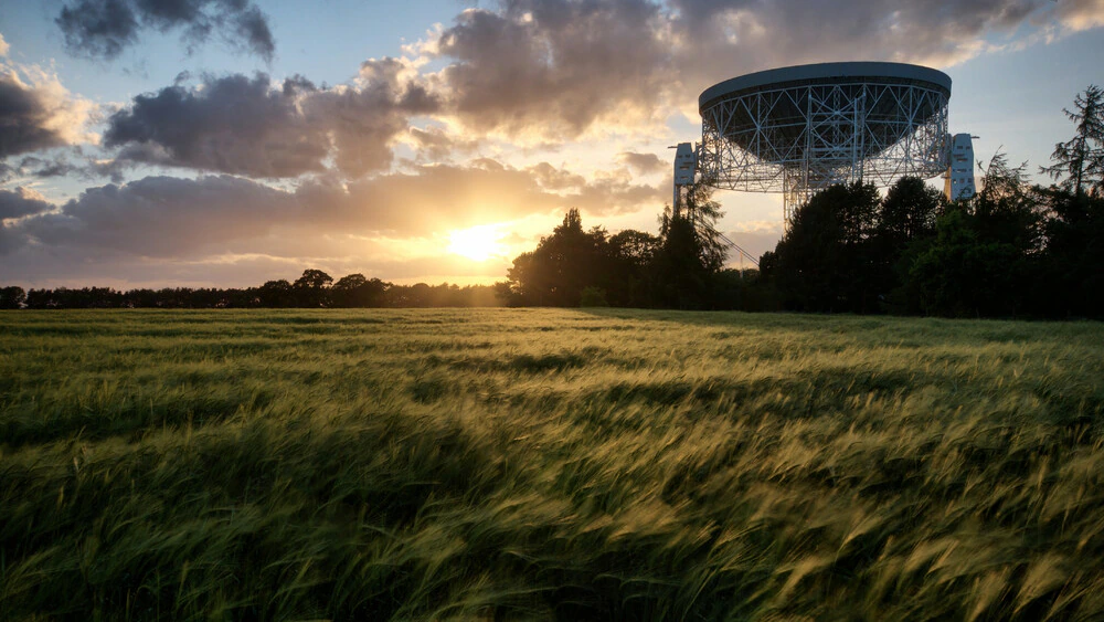 A radio telescope rises above a grassy field at sunset, with clouds and trees in the background as sunlight streams across the landscape.
