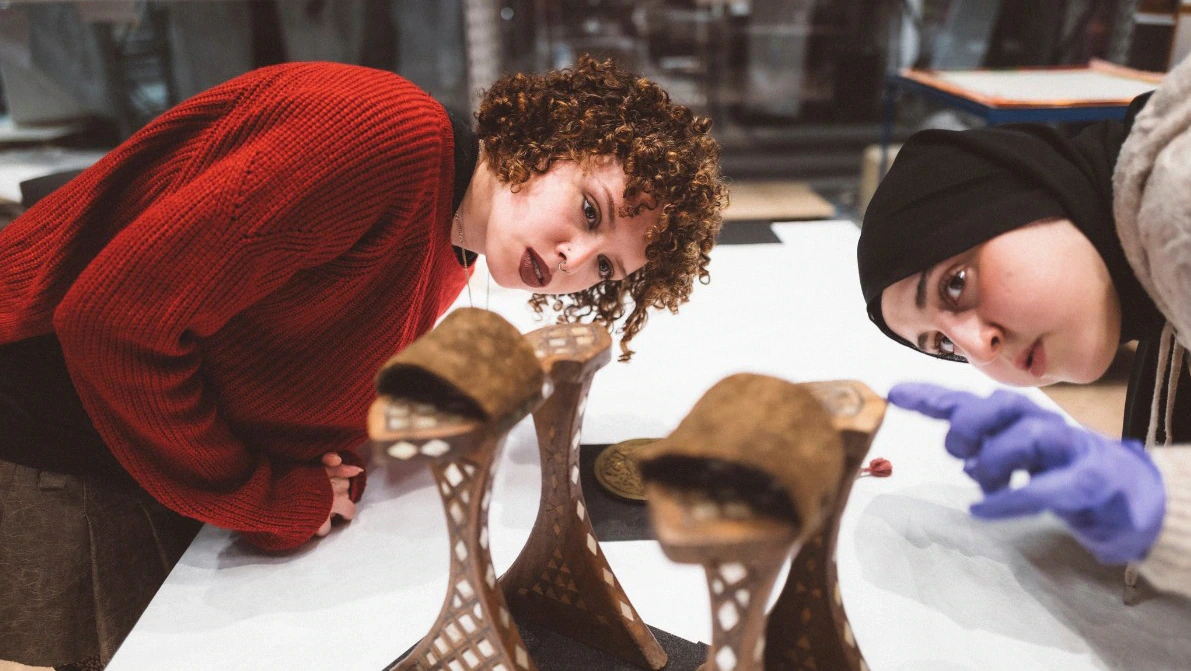 Two women closely examine intricate architectural models on a table; one wears a red sweater, the other a black hijab and blue gloves. The models feature curved structures with lattice-like bases.