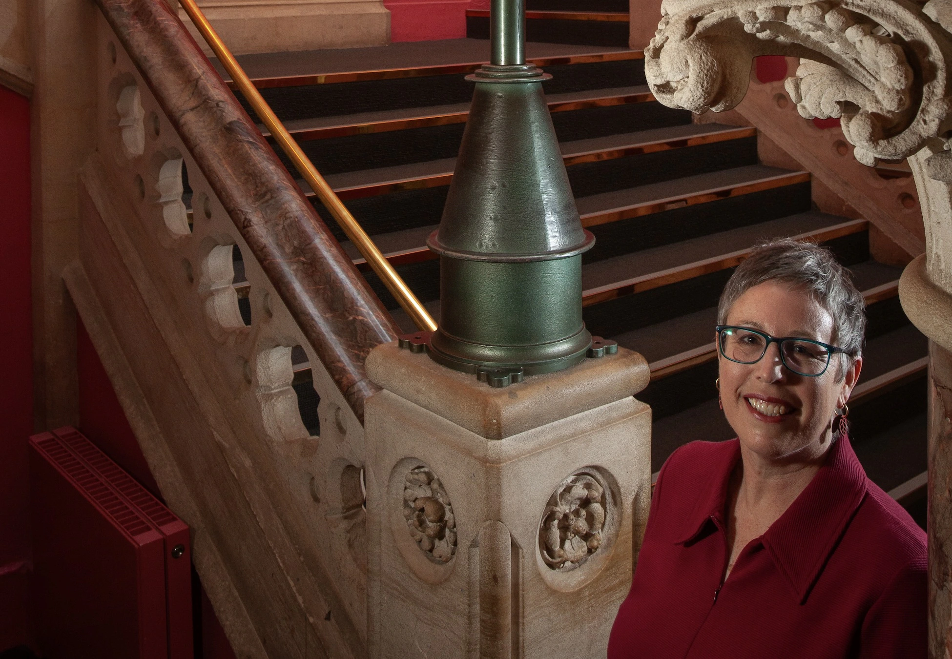 A woman with short gray hair and glasses, wearing a red top, stands smiling near an ornate stone staircase with intricate railings and decorative columns.