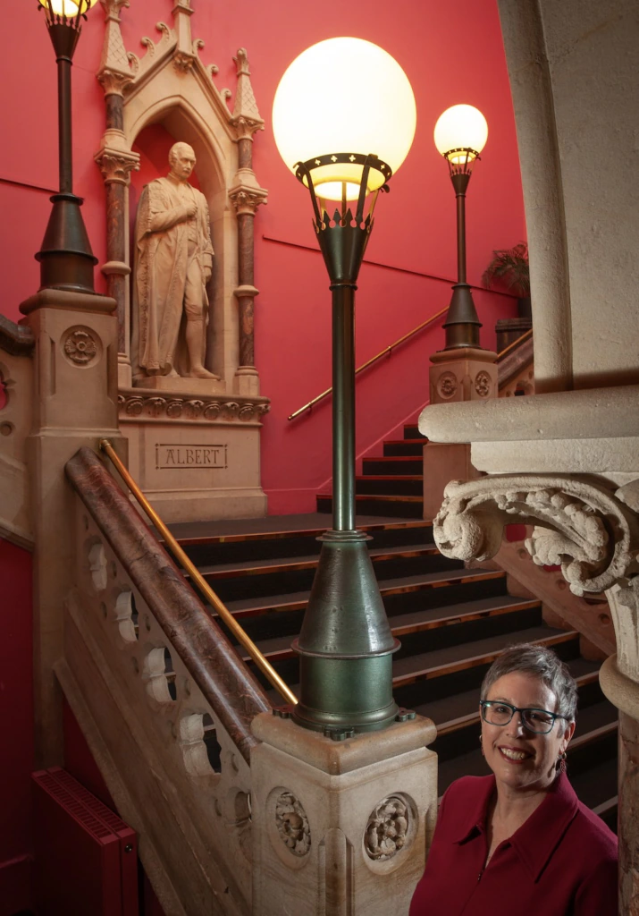 A smiling woman in glasses stands by ornate stairs with a statue labeled Albert and vintage lamps against red walls in the background.