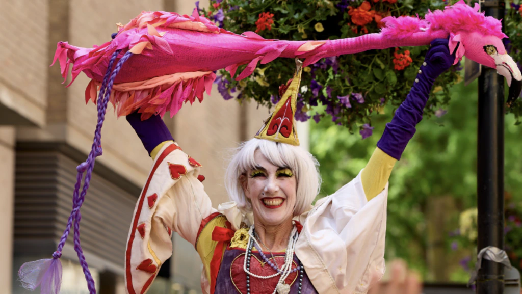 A performer in colorful costume and hat holds a large pink bird puppet overhead, smiling, with flowers and greenery in the background.