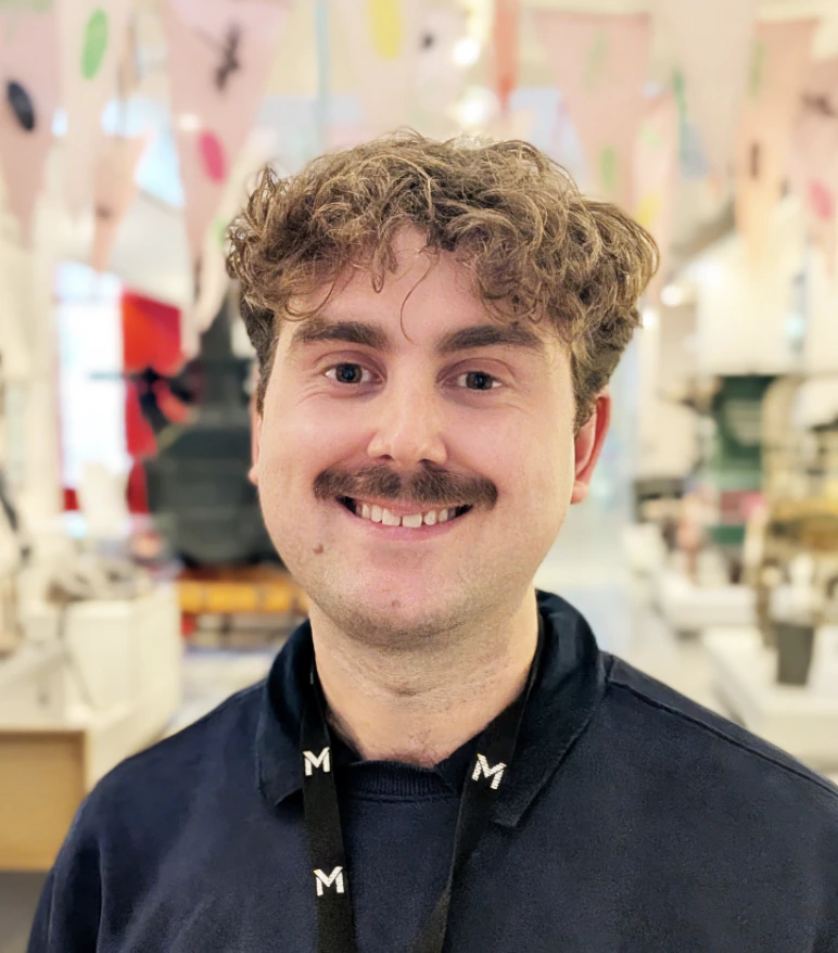 A smiling person with curly brown hair and a mustache wears a black lanyard with an M on it. Colorful banners and shelves are visible in the bright background.