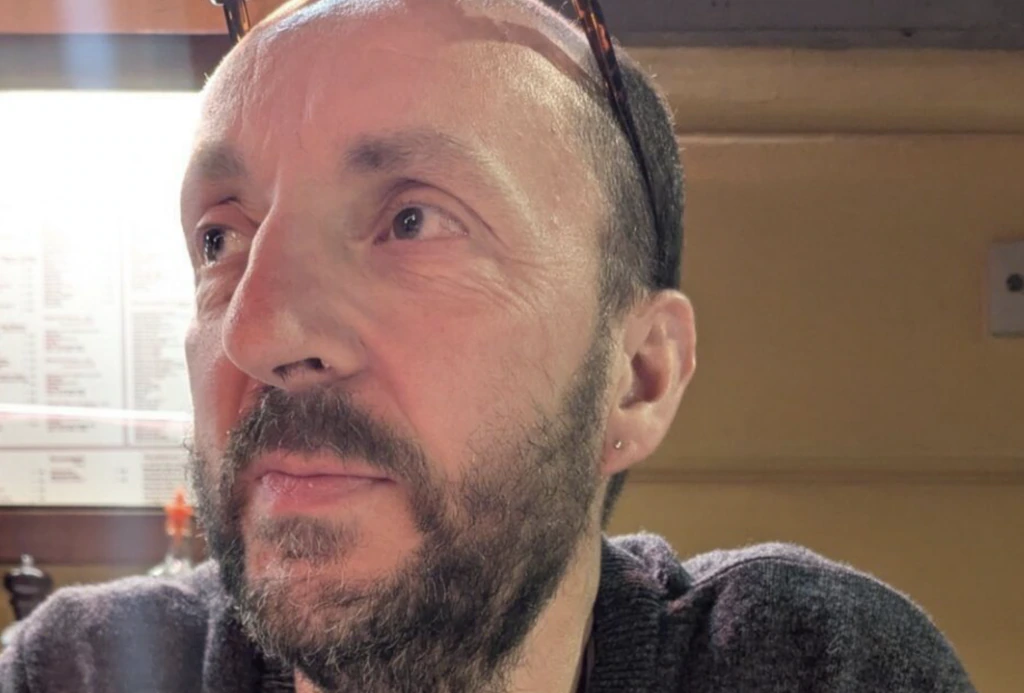 A man with a beard and short hair sits indoors. He wears glasses on his head and a dark sweater. The background shows a menu and warm lighting.