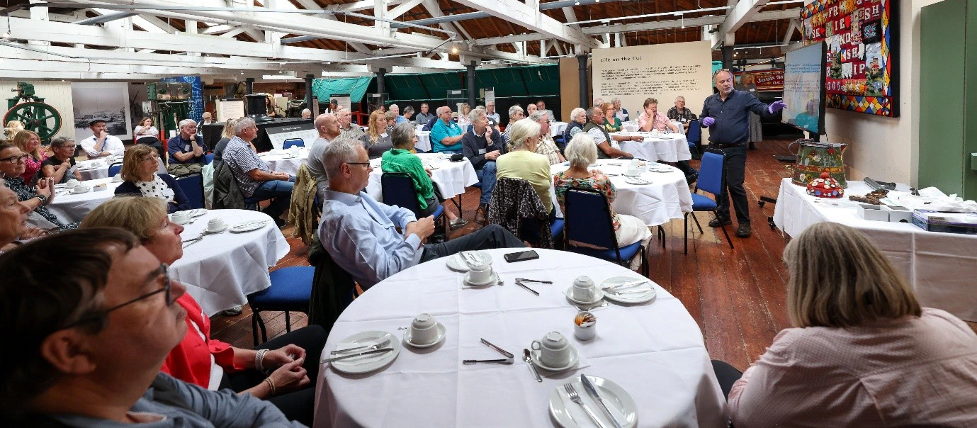 A group of people sits at round tables in a bright hall, listening to a speaker standing near a quilt on the wall. Tea cups, plates, and cutlery are set on the tables. The ceiling has wooden beams.
