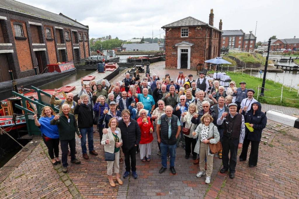 A large group of people poses together outside at a historic canal site, surrounded by brick buildings and boats, many smiling and raising glasses, on a cloudy day.