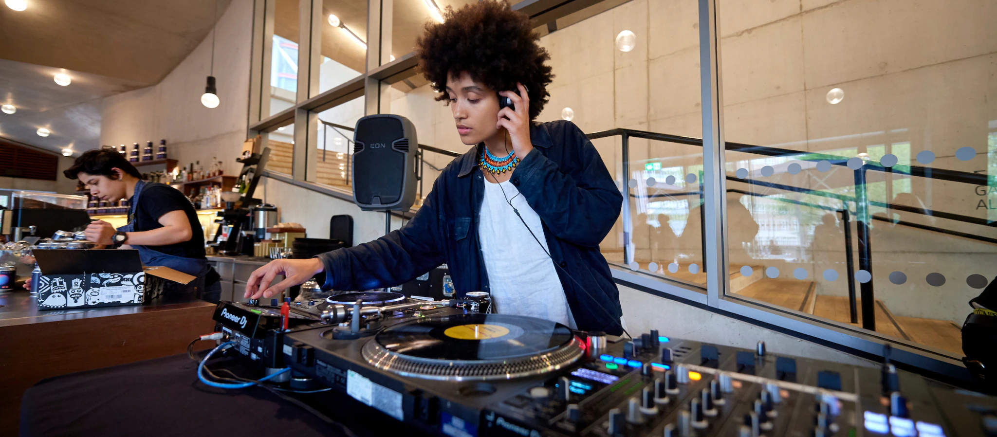 A DJ with curly hair mixes vinyl records at a turntable setup indoors, wearing headphones and a necklace. Another person works in the background near a bar area with shelves and hanging light bulbs.