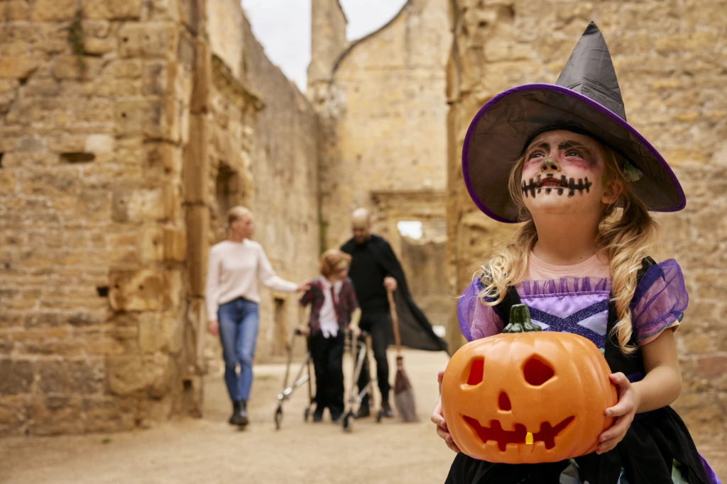 A young girl in a witch costume and face paint holds a pumpkin bucket, standing in front of old stone ruins. Three adults in Halloween costumes are blurred in the background.