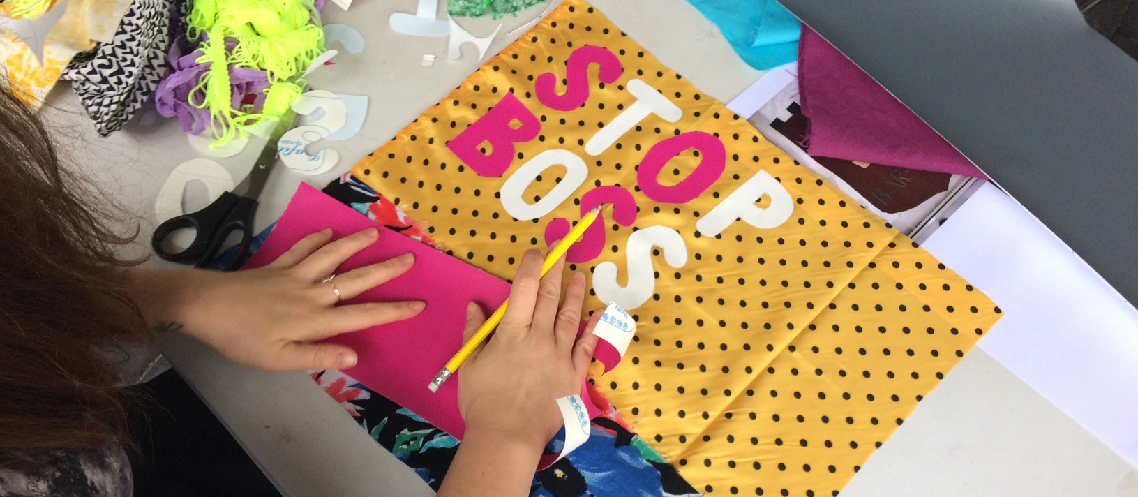 A person traces letters onto pink paper at a desk, working on a colorful sign with fabric that reads STOP BOSS in large white letters. Craft materials and fabric pieces are scattered around.