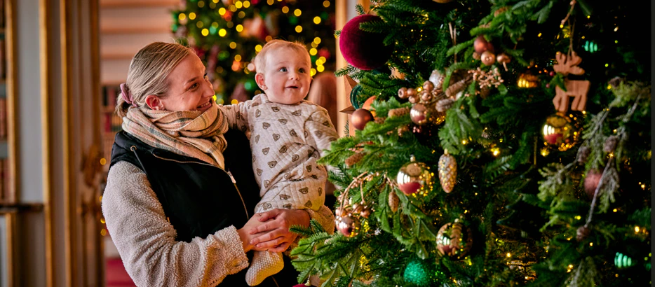 A woman holding a baby stands beside a decorated Christmas tree with gold, red, and green ornaments and twinkling lights, both admiring the festive decorations.