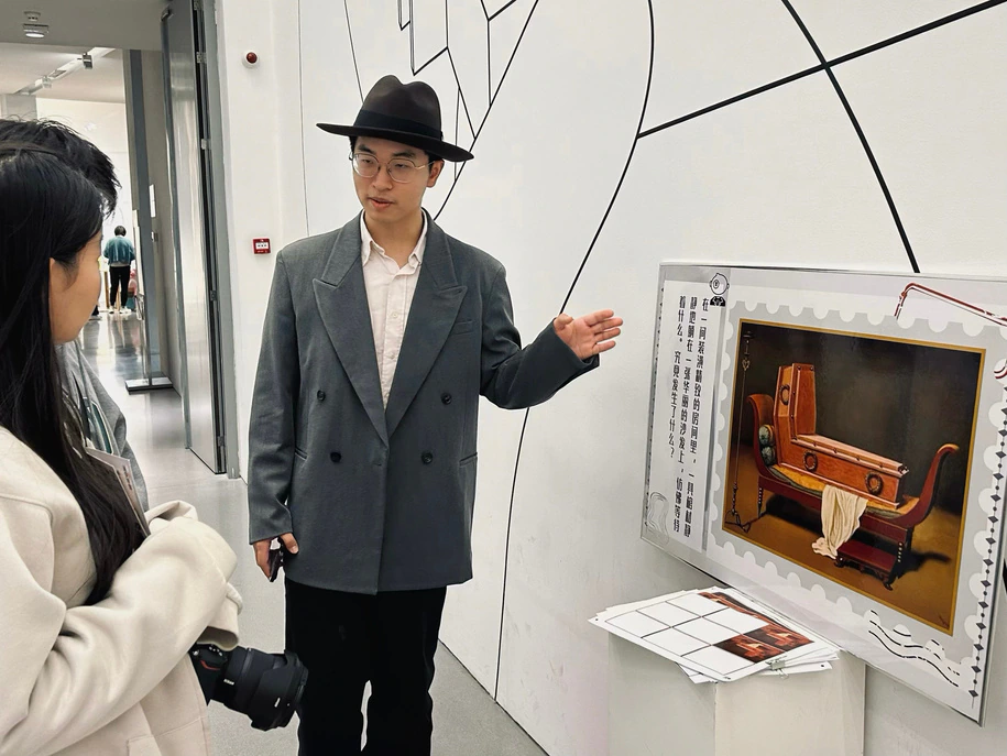 A person wearing a hat and glasses gestures toward a framed artwork on display, while another person listens. The scene takes place indoors with modern decor and white walls.