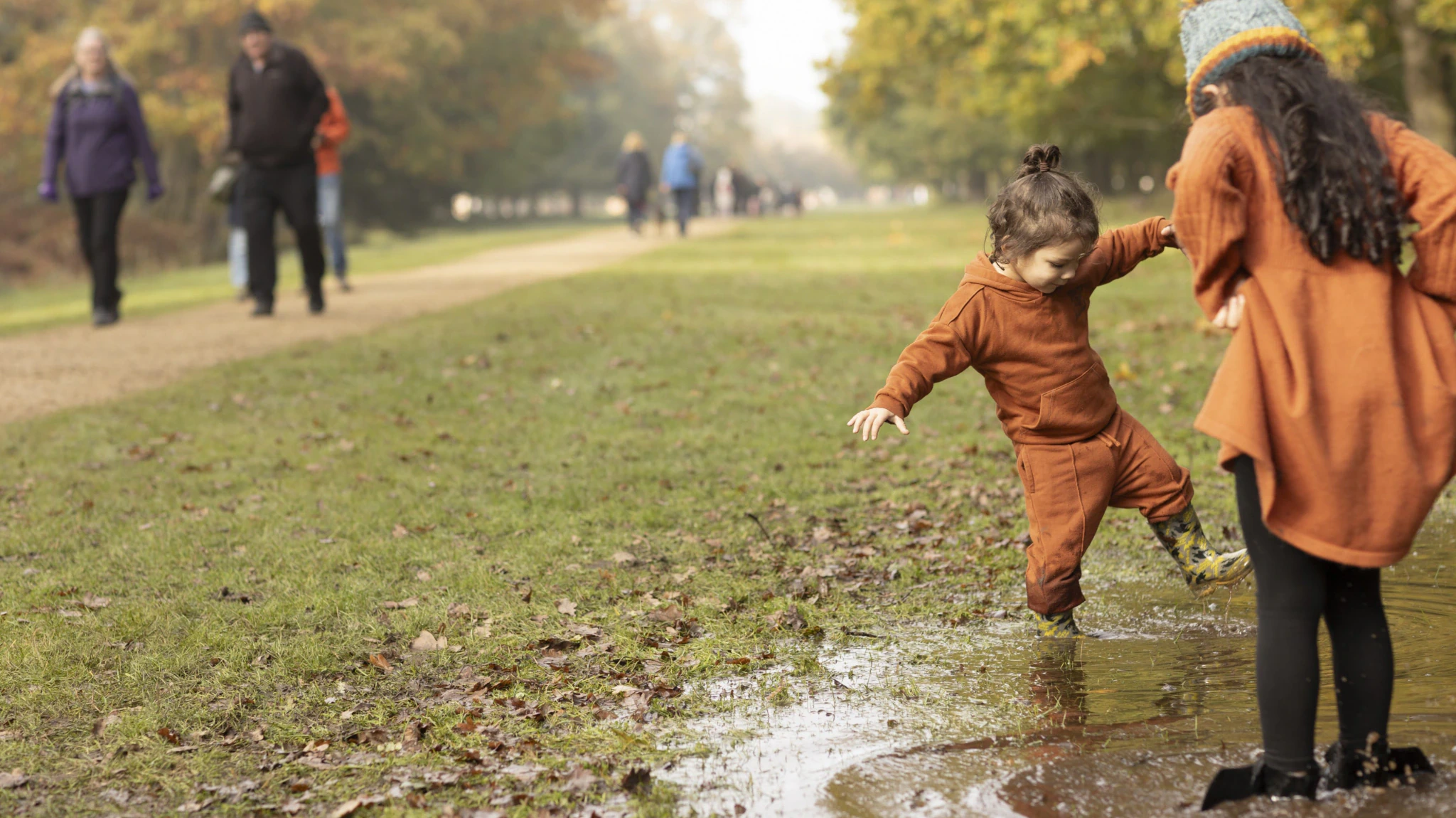 Two children in autumn clothes splash in a puddle on a grassy park path, while several adults walk in the background among trees with yellow and orange leaves.