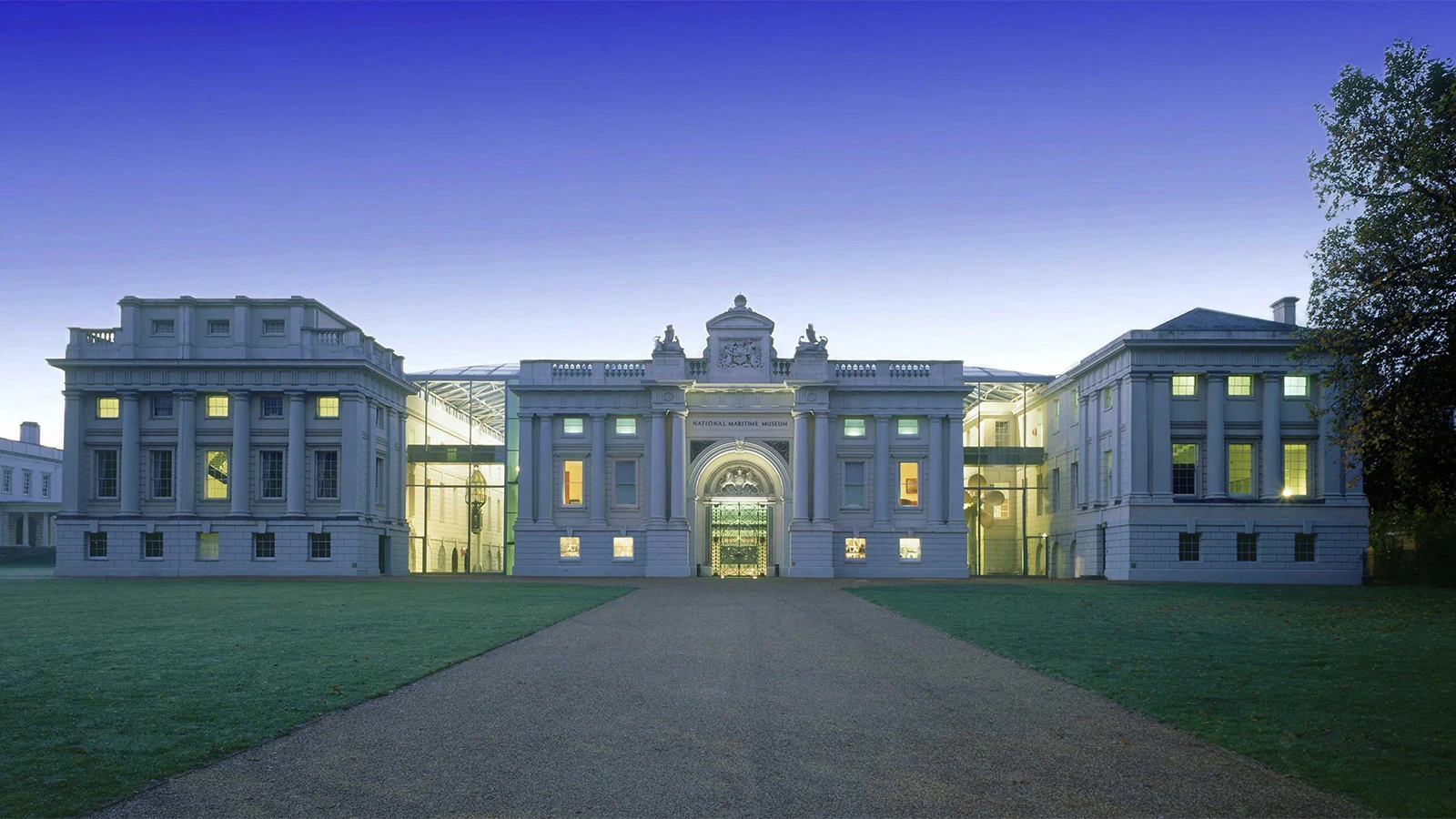 A grand neoclassical building with large windows is illuminated at dusk, featuring a central arched entrance, a decorative pediment, and symmetrical wings on both sides, all set against a vivid blue sky.