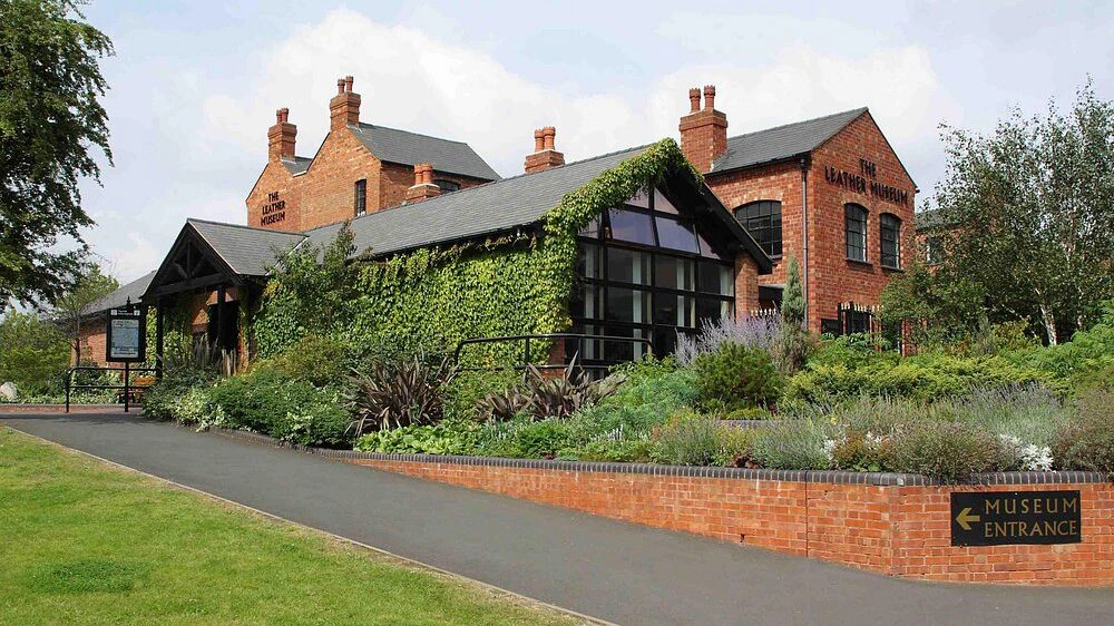 Red brick museum building covered partly with ivy, surrounded by greenery and landscaped plants, with a sign pointing left toward the entrance.