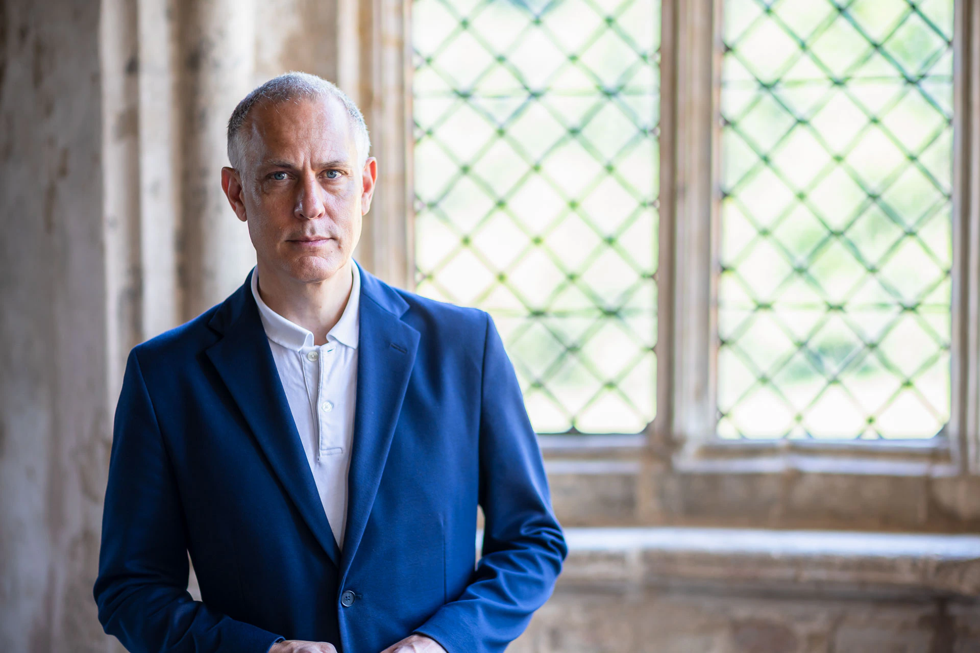 A man in a blue blazer and white shirt stands indoors near a window with diamond-patterned glass, looking directly at the camera.