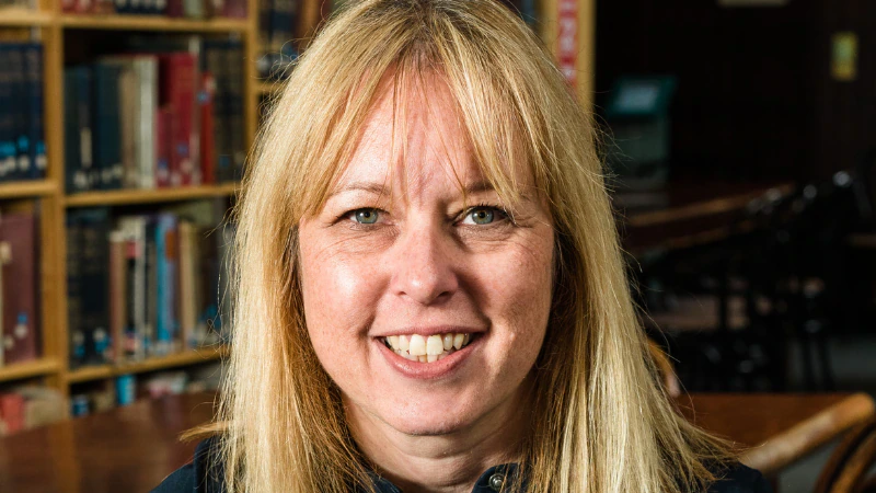 A smiling woman with blonde hair sits at a wooden table in a library, with bookshelves in the background.