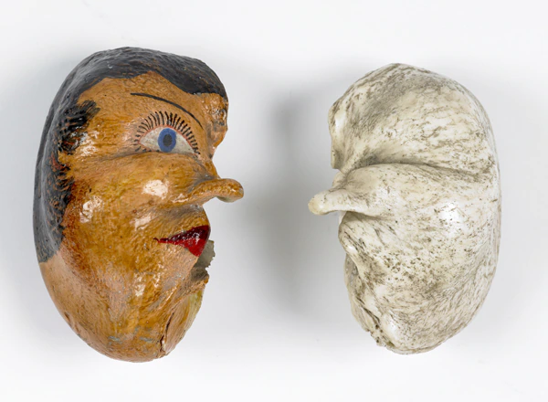 Two objects shaped like beans: one painted with a human face, featuring an eye, lips, and black hair; the other unpainted, white, and textured, resembling a natural bean. Both are positioned side by side on a white background.