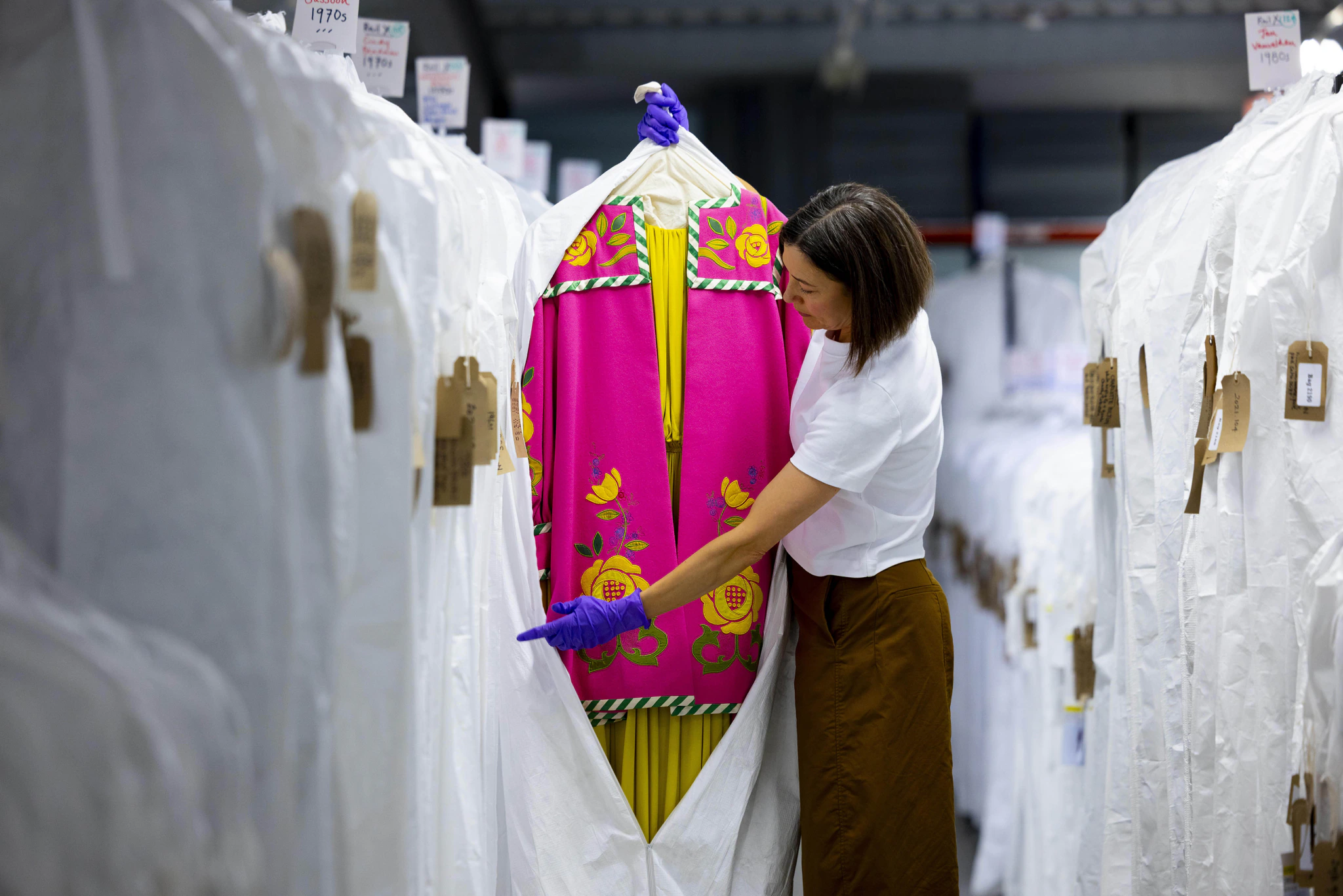 A person wearing gloves examines a bright pink and yellow embroidered garment stored in a protective cover among rows of covered clothes on racks.