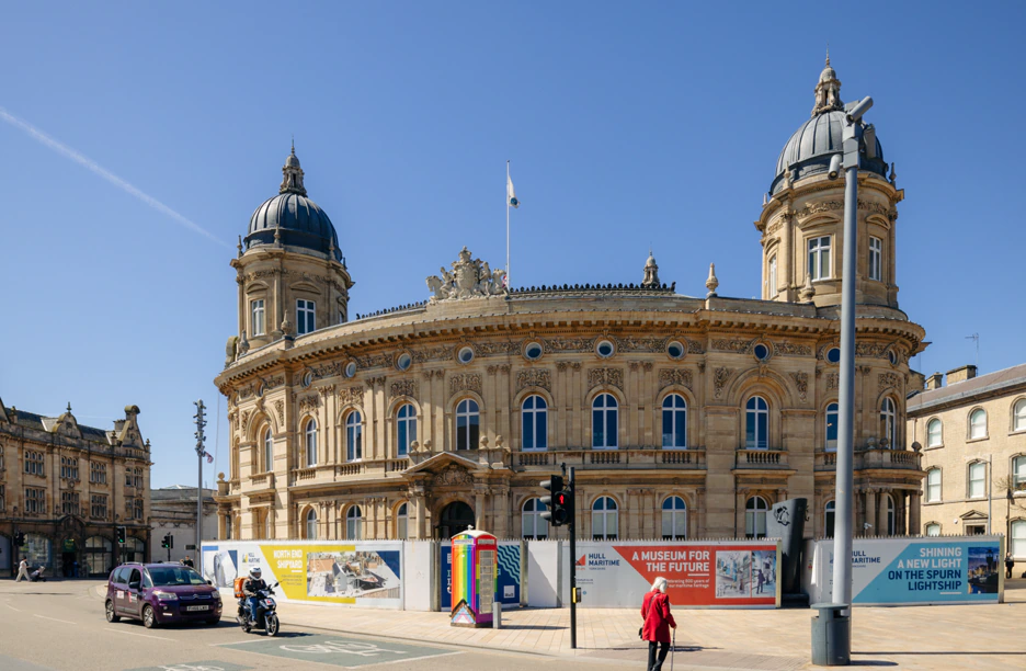 Historic domed building with ornate stonework, flagpole on top, and temporary construction barriers featuring colorful signs in front. Clear blue sky. People, a motorbike, and a traffic light are visible in the foreground.