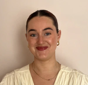 A woman with dark hair pulled back, wearing gold hoop earrings, a gold chain necklace, and a cream-colored top, smiles at the camera against a plain, light background.