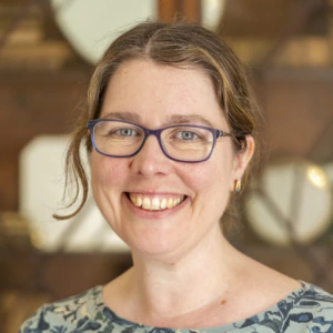 A woman with glasses and a patterned shirt smiles, standing indoors in front of a wooden cabinet with glass doors and shelves containing books and decorative objects.