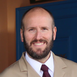 A man with a beard and bald head, wearing a tan suit and tie, stands and smiles near a green doorframe with a blue door and signs in the background.