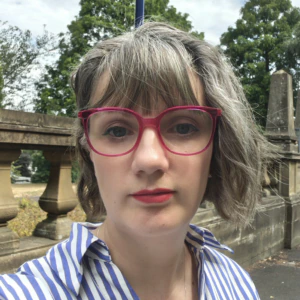 A person with short, wavy brown and gray hair, wearing red glasses and a blue striped shirt, stands outside in front of a stone fence and trees on a cloudy day.