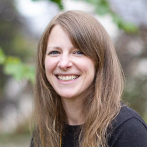 A woman with long brown hair and a black top smiles outdoors, with blurred green leaves and trees in the background.