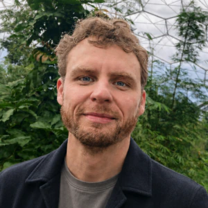 A man with light brown hair and a beard stands in front of lush green plants inside a large geodesic greenhouse structure.