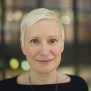 A person with short blonde hair, light skin, and blue eyes wears a black top and a red beaded necklace, standing in front of a blurred background with warm lighting.