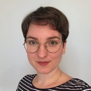 A person with short brown hair, wearing round glasses and a black-and-white striped shirt, stands in front of a plain light background, looking at the camera and smiling slightly.