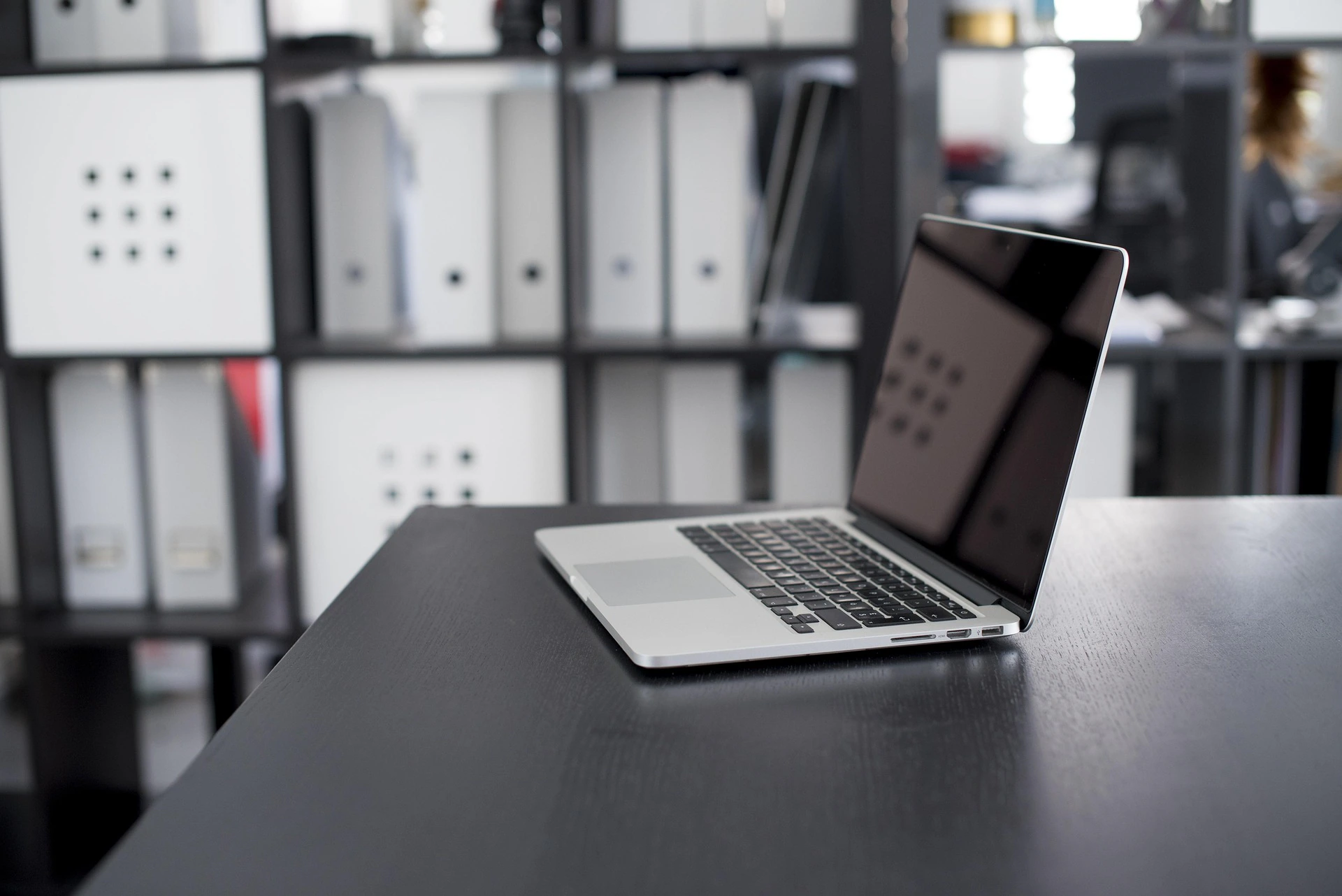A silver laptop sits open on a dark desk in an office with shelves filled with white and gray storage boxes in the background.
