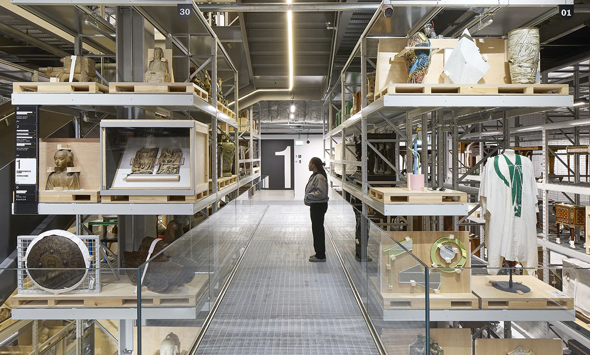 A person stands on a walkway in a museum storage area, surrounded by shelves filled with sculptures, artifacts, and textiles. The space is industrial with metal racks and bright lighting.