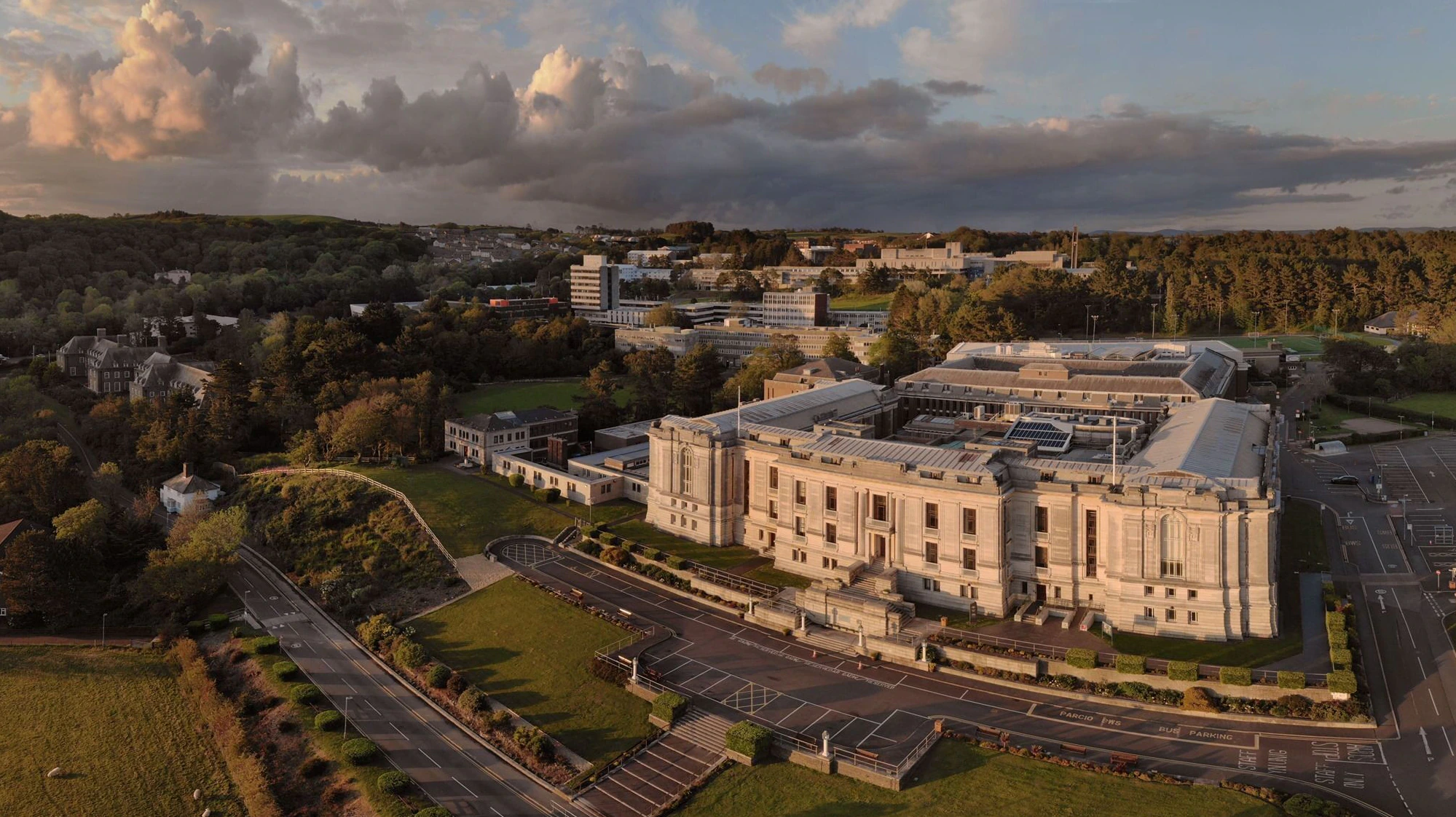 A large, historic building surrounded by trees and modern structures, with dramatic clouds in the sky and sunlight casting long shadows across the landscape.