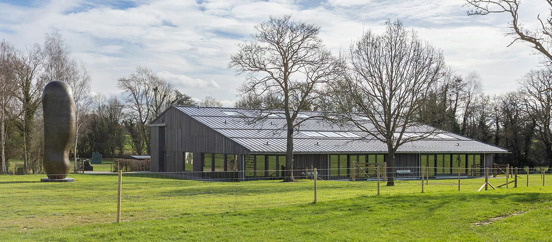 A modern building with large windows sits on a green lawn, surrounded by trees. A tall, abstract sculpture stands to the left under a partly cloudy sky.