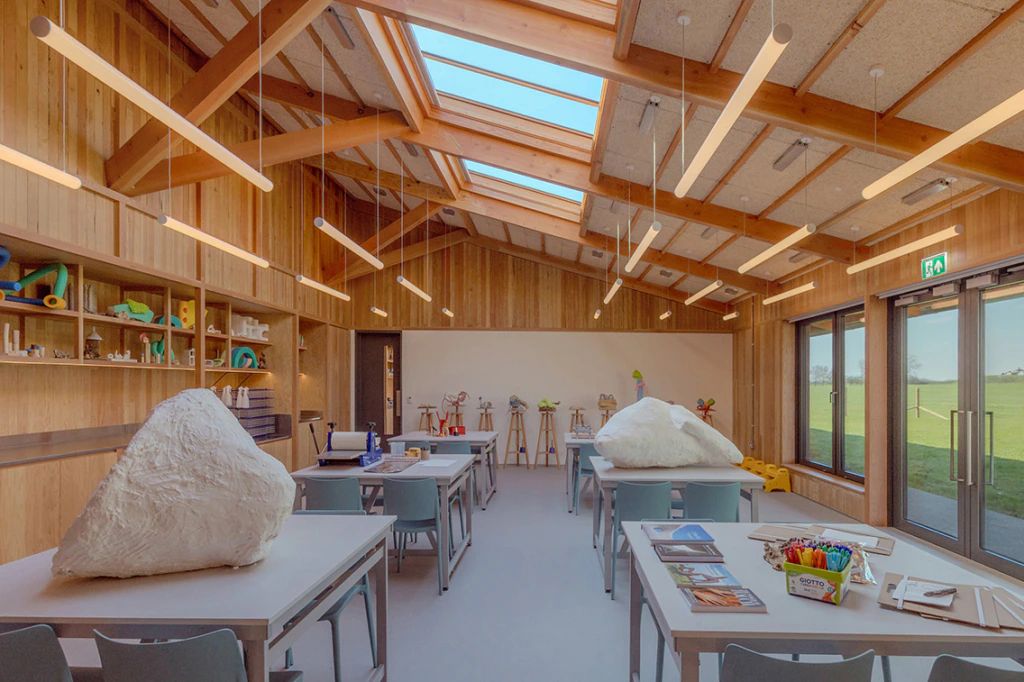 A bright art classroom with wooden walls, tables, art supplies, and sculptures. Large windows and skylights let in natural light, and fields are visible outside.