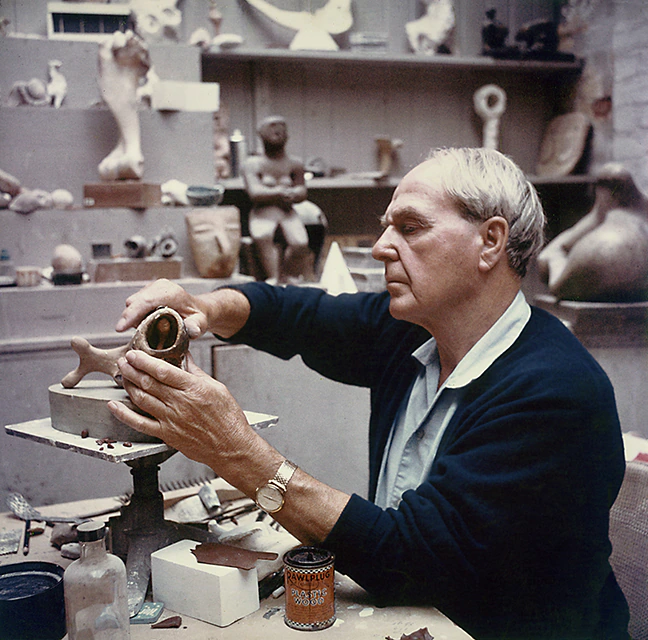 A man shapes a clay sculpture in a studio filled with various artworks and tools. He is focused on his work, wearing a dark jacket and gold watch, surrounded by jars and finished sculptures.