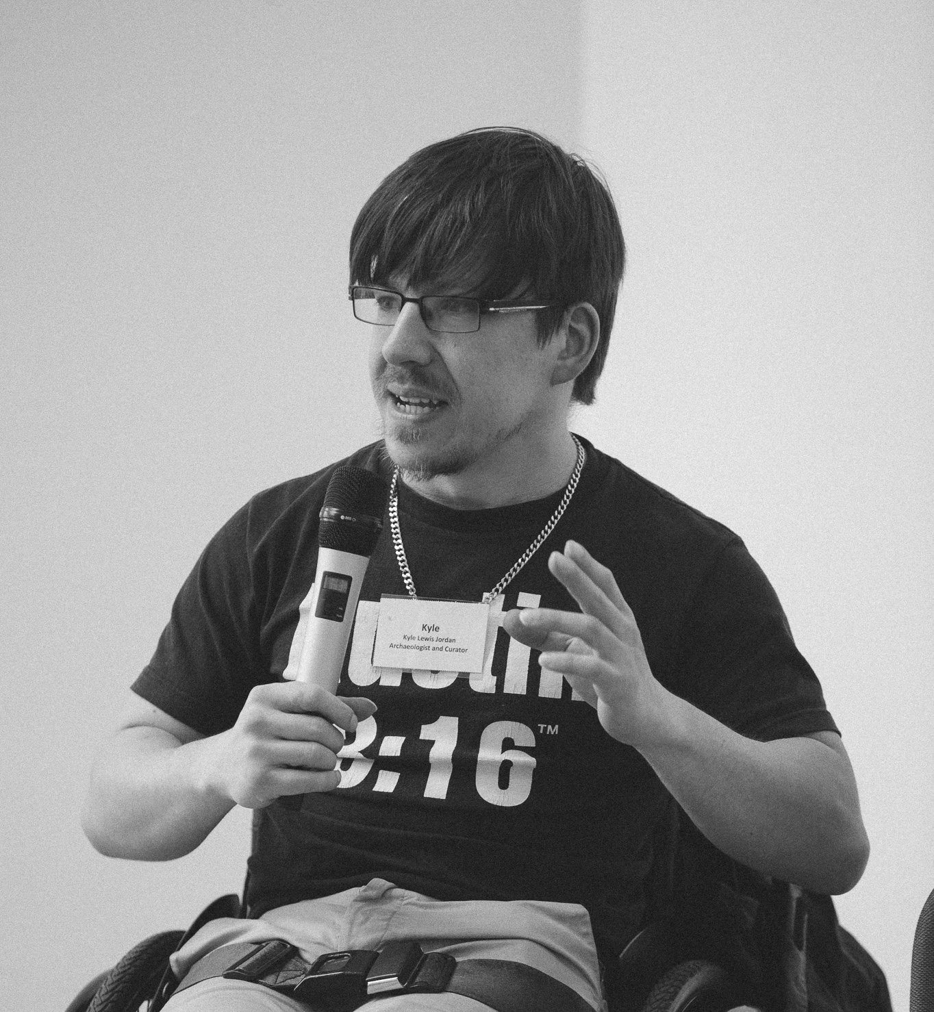 A person in a wheelchair speaks into a microphone while gesturing with their hand. They wear glasses and a t-shirt with a visible name tag and text. A water bottle and glasses are on the table in front of them.