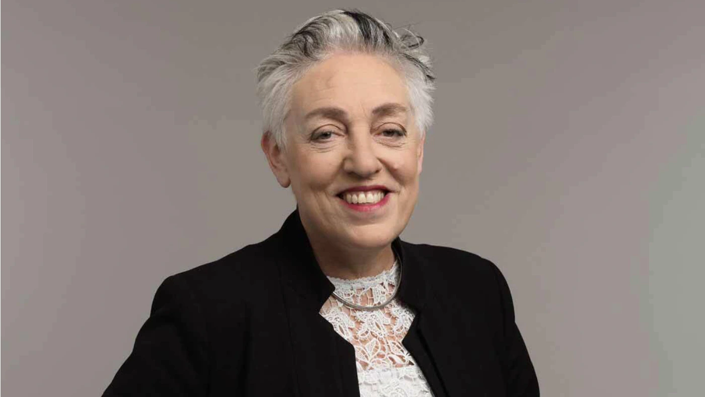 Smiling person with short gray hair wearing a black blazer and a white lace top, standing against a plain light gray background.