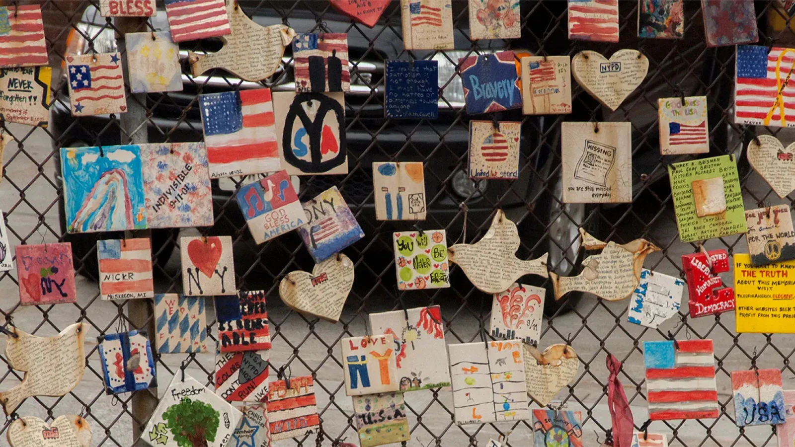 Handmade tiles with patriotic and heartfelt messages hang on a chain-link fence, featuring American flags, peace signs, hearts, and handwritten notes.