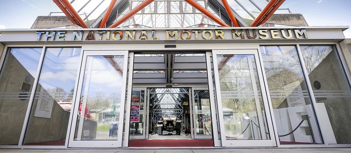 The entrance of The National Motor Museum with glass doors and a geometric glass roof above, sunlight streaming in, and museum signs visible.