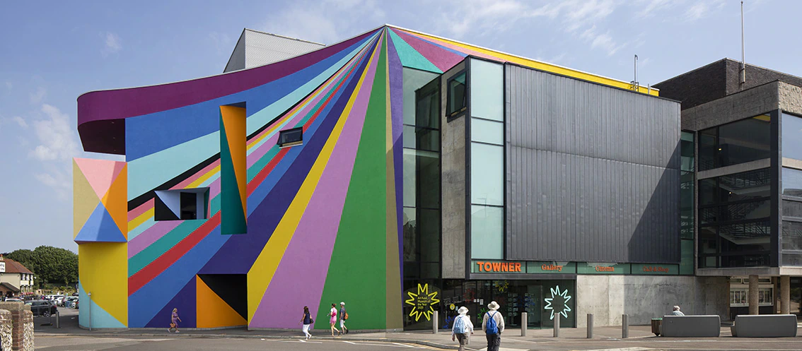 A modern building with large colorful geometric mural, featuring bold stripes and triangular patterns. Several people walk nearby, and the sky is mostly clear with a few clouds.
