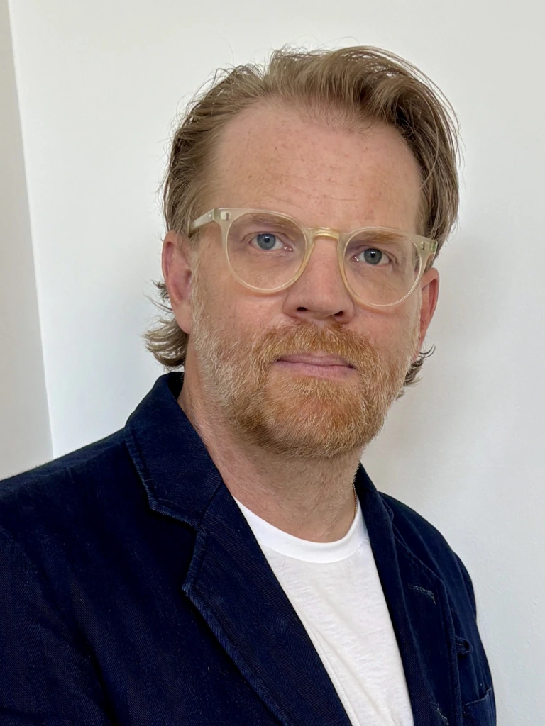 A man with light skin, short light brown hair, beard, and clear-framed glasses wears a navy jacket over a white shirt, standing against a plain white background.